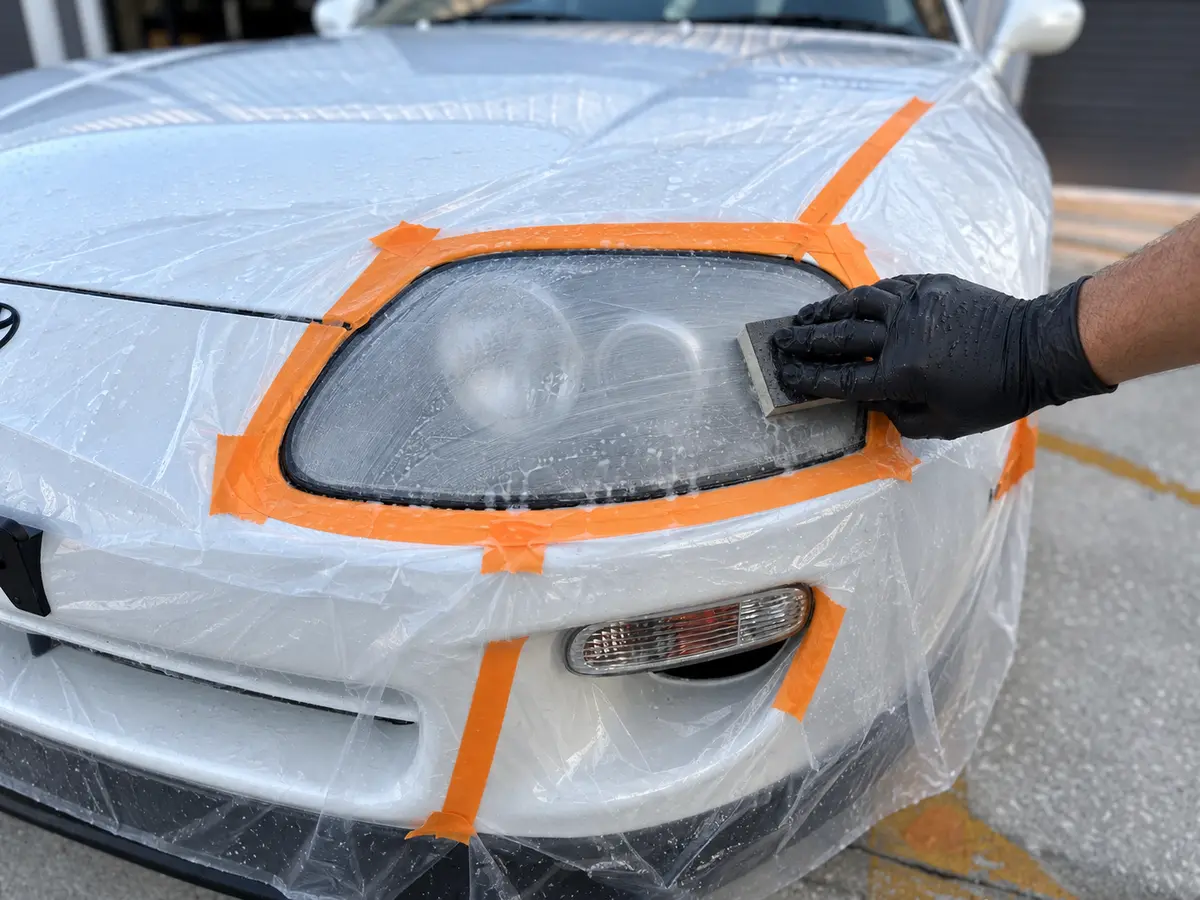 White Toyota Supra headlight undergoing multi-stage wet sanding to remove failed coating and oxidation